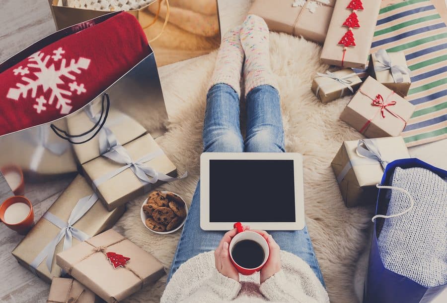 Image of woman sitting on the floor with a tablet. She is surrounded by wrapped boxes and gift bags