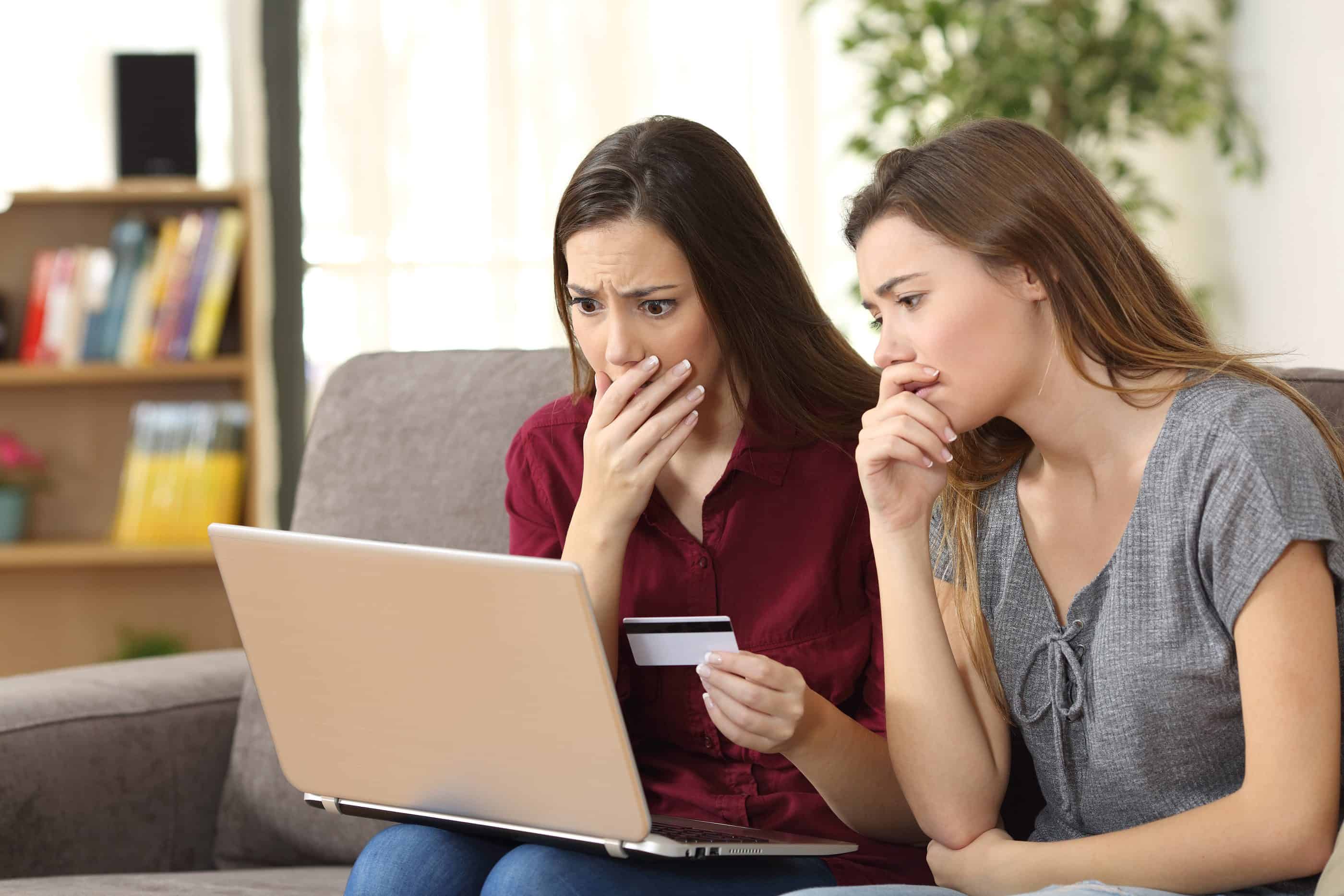Two worried friends having problems buying on line with credit card and a laptop sitting on a couch in the living room at home