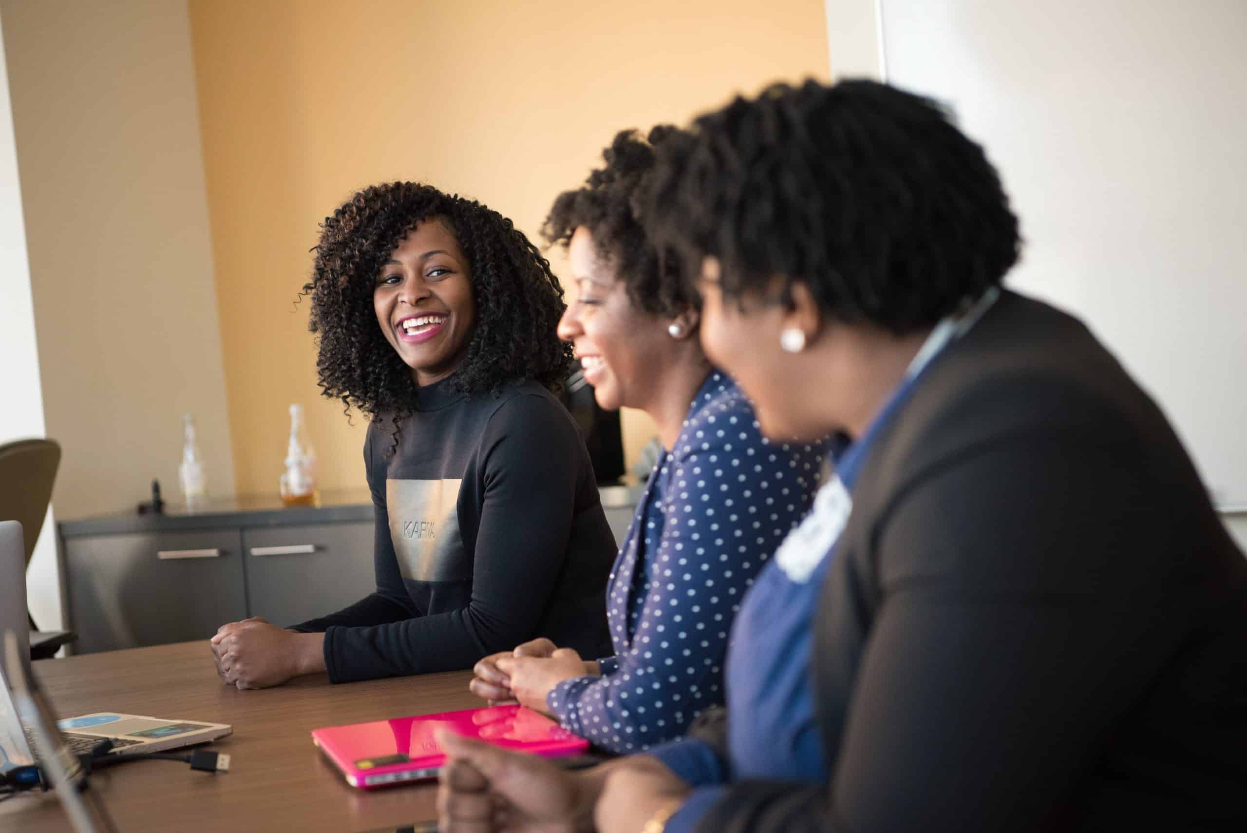 Photo of three feminine-presenting people of color smiling and talking at a table in an office space. They are in business casual clothing.